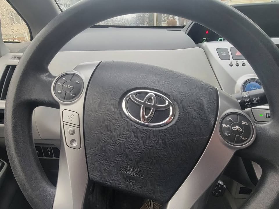 Close-up of steering wheel in a 2015 Toyota Prius v, showing audio, trip, and temperature controls with dashboard in background