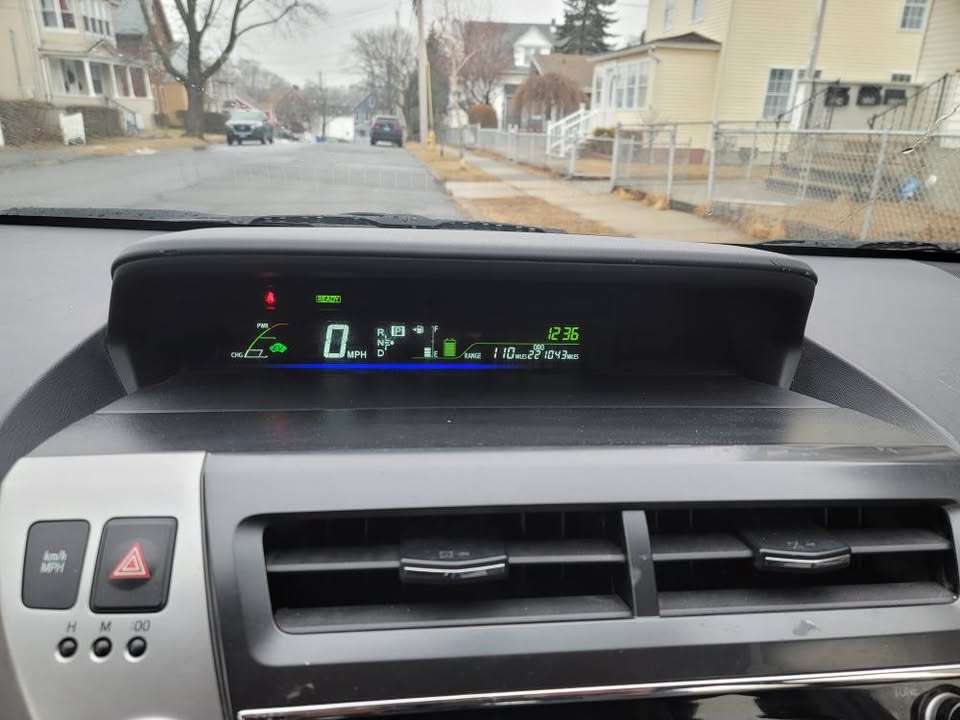 Wide view of 2015 Toyota Prius v dashboard showing 110-mile range, 210,043 miles on the odometer, 0 MPH, and a clear digital speedometer in a residential neighborhood