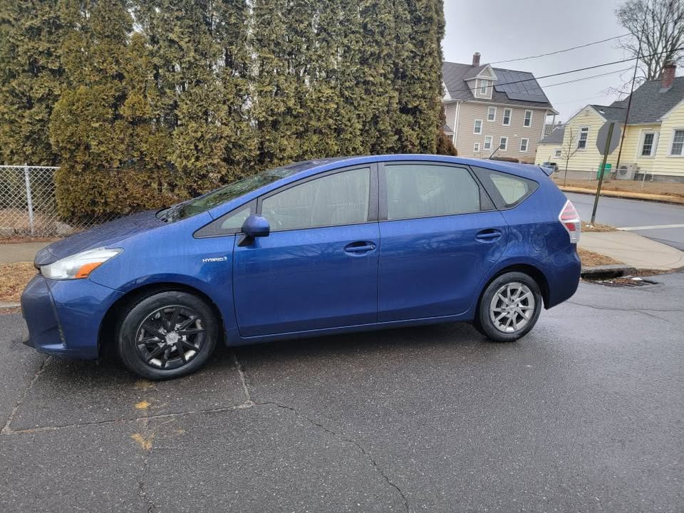 Side profile of a blue 2015 Toyota Prius v Base parked on a wet street near residential homes and tall hedges