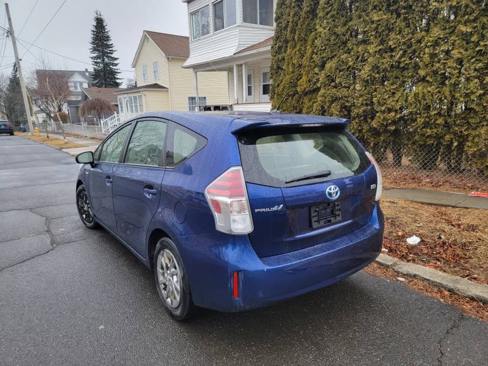 Rear and side view of a blue 2015 Toyota Prius v parked on a residential street, showing taillights, hatchback design, and overcast weather setting