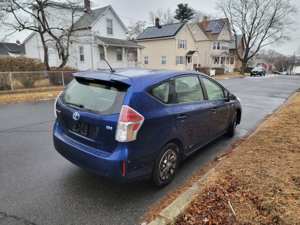 Angled rear view of a blue 2015 Toyota Prius v parked on a wet street in a suburban neighborhood, showcasing hatchback, taillights, and body condition
