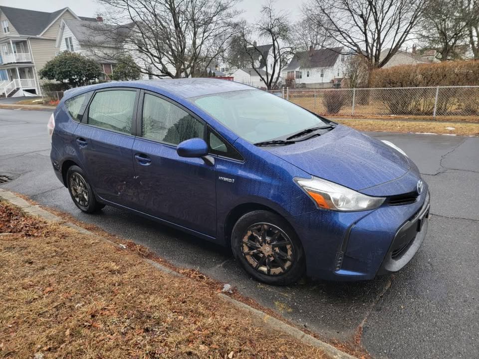 Front-side view of a blue 2015 Toyota Prius v parked curbside on a rainy day, showing driver's side doors, hybrid badge, and headlight detail