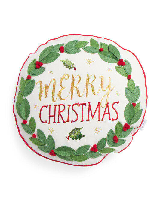A round white throw pillow with a red and green Merry Christmas wreath design, featuring metallic stitching and floral applique.