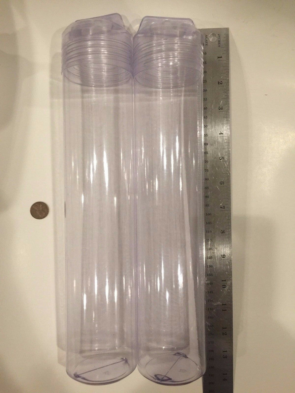 Two tall clear plastic tubes with a ruler next to them on a plain background.