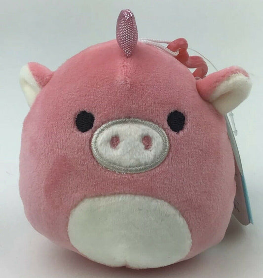 A plush toy resembling a pink unicorn with a clip-on feature, has black eyes, a white nose, and a tuft of hair on the top. It is round in shape and has a tag attached.