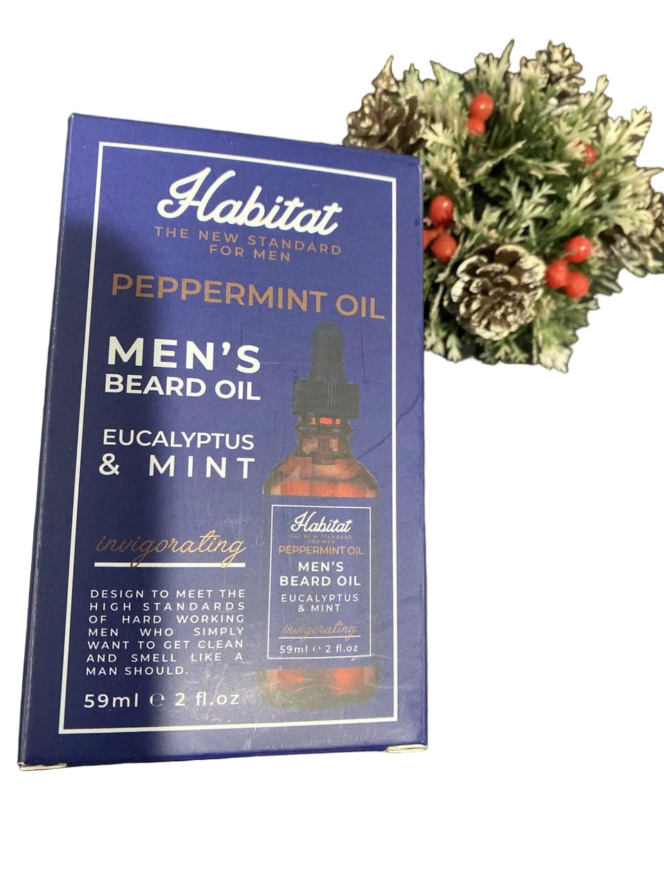 A bottle of Home & Body Company Habitat Peppermint Oil Men's Beard Oil with packaging that includes the brand name, product name, and a depiction of the bottle. The packaging is displayed in a setting with decorative items in the background.