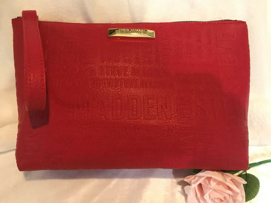 A red Steve Madden T-bottom cosmetic bag with a gold zipper and logo, displayed on a white surface with a pink flower to the side.