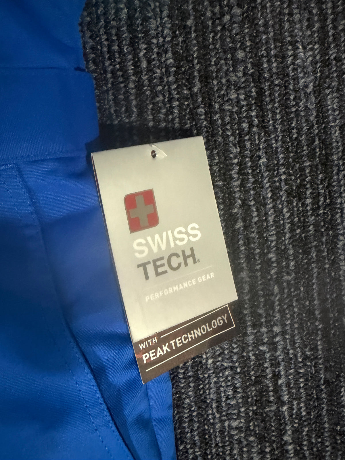 A close-up of a blue garment with a silver and black 'SWISS TECH' hangtag on a dark gray textured background.