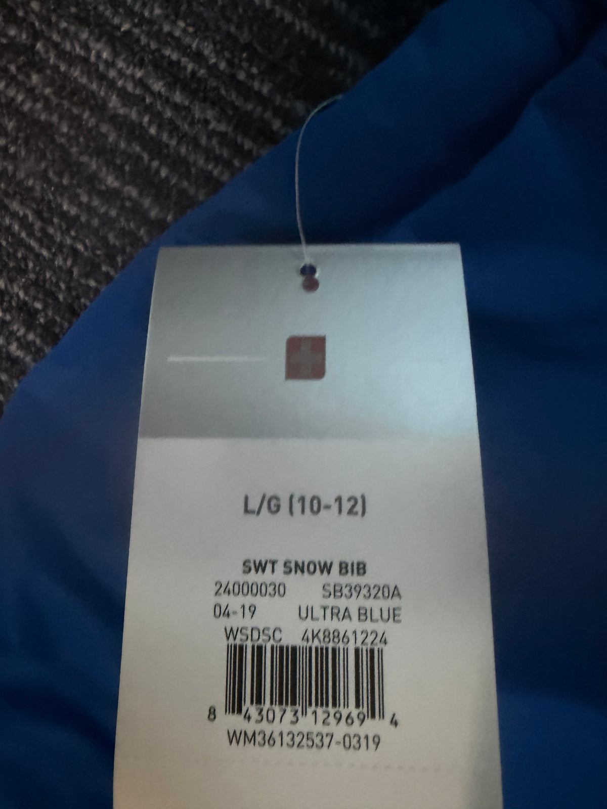 A close-up of a white garment tag attached to a blue fabric item, showing printed text including 'SWT SNOW BIB' and a barcode 