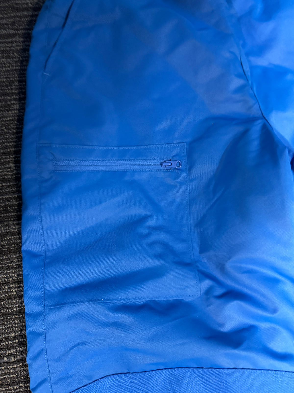 A close-up of a bright blue snow pants  showing a zippered chest pocket and fabric texture, laid flat on a dark textured surface.
