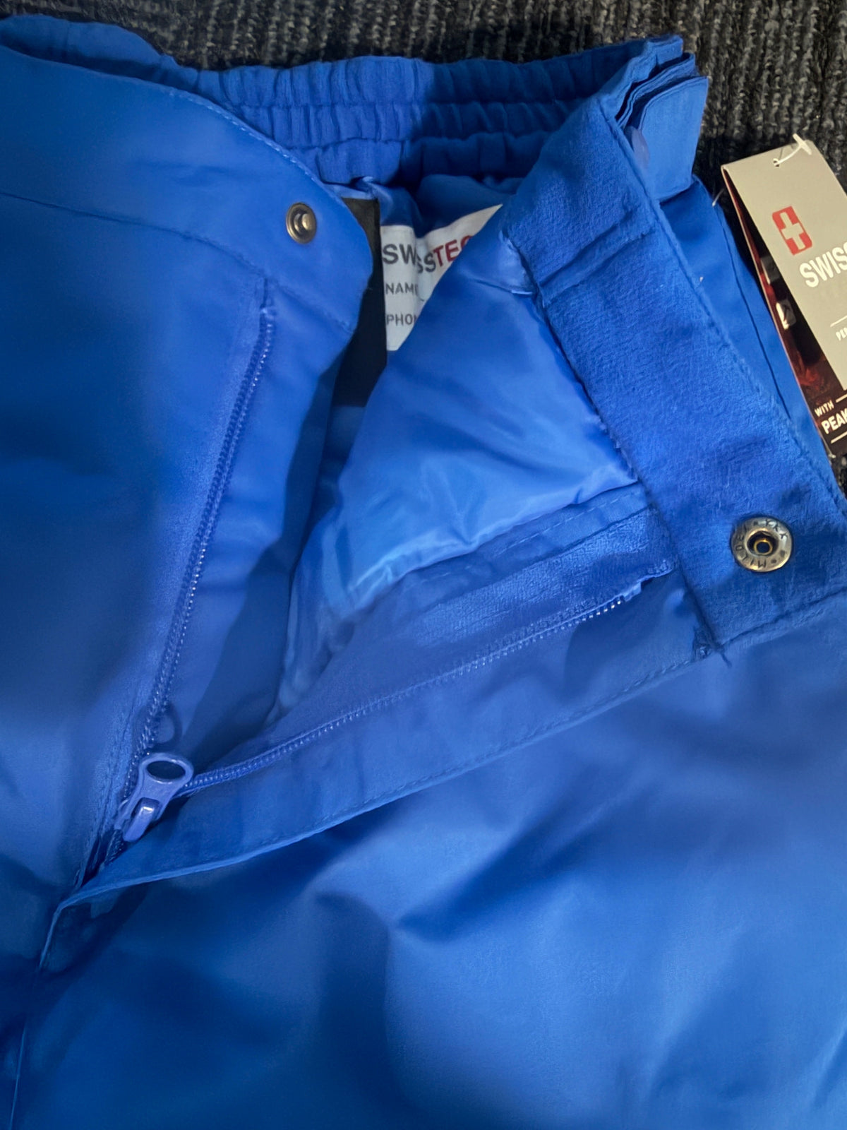 A close-up of a bright blue snow pants outerwear piece, showing the waistband, zipper, snap button, and part of the interior lining. A hangtag is visible at the top edge.