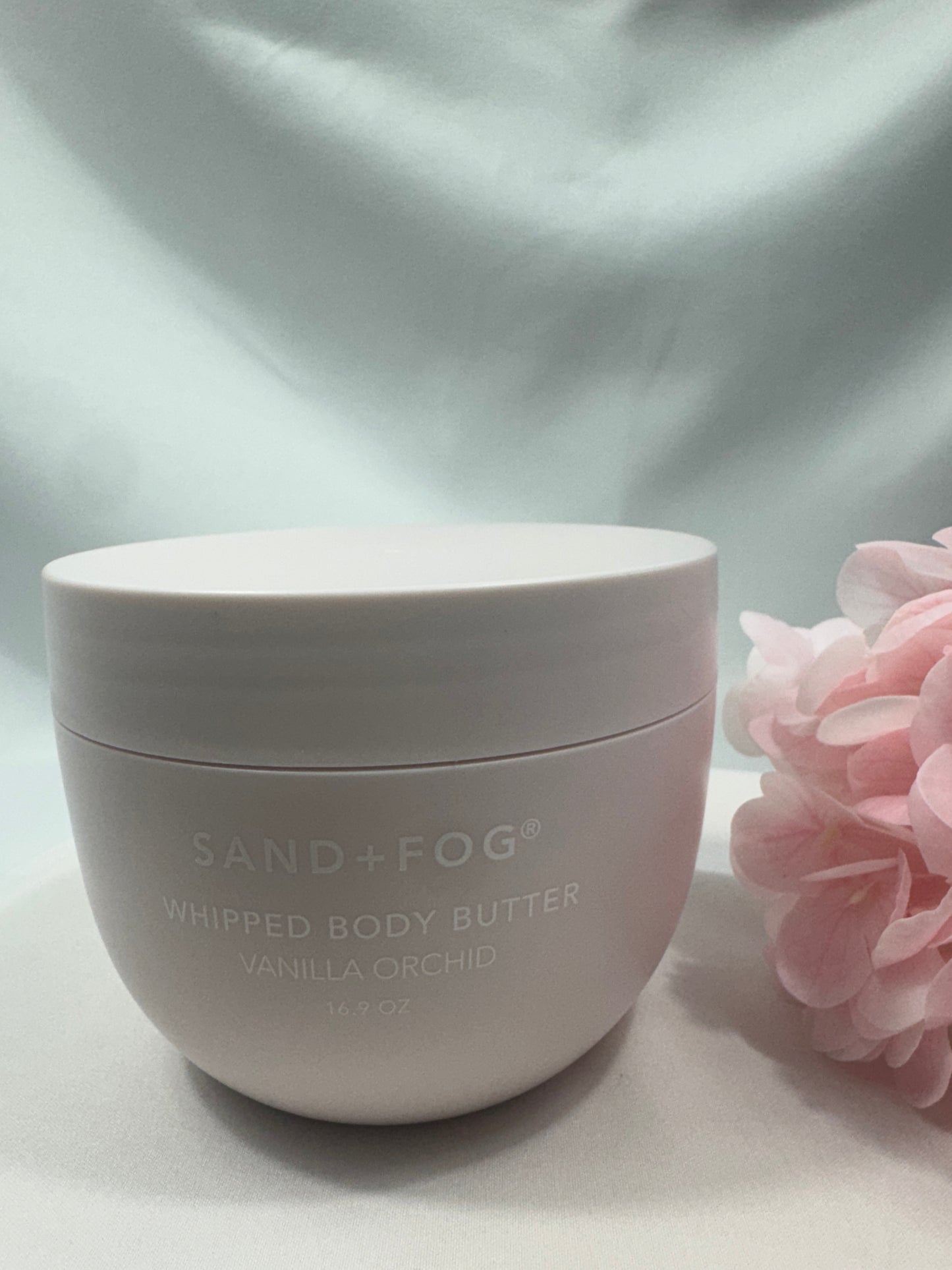 White container of Sand + Fog whipped body butter with pink flowers in the background