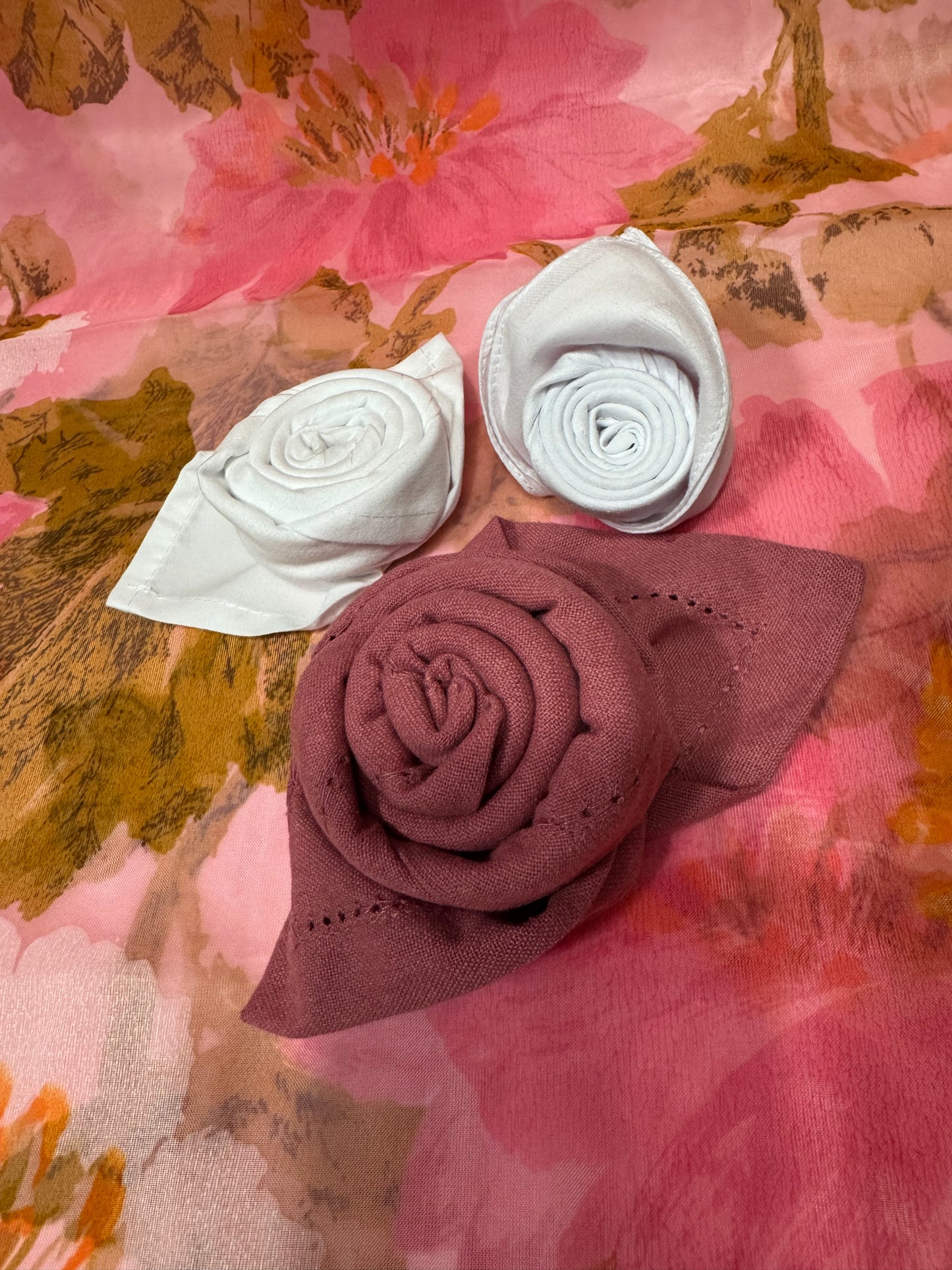 Three rolled cotton napkins, two in white and one in burgundy, shaped into rose-like folds, placed on a colorful floral fabric background