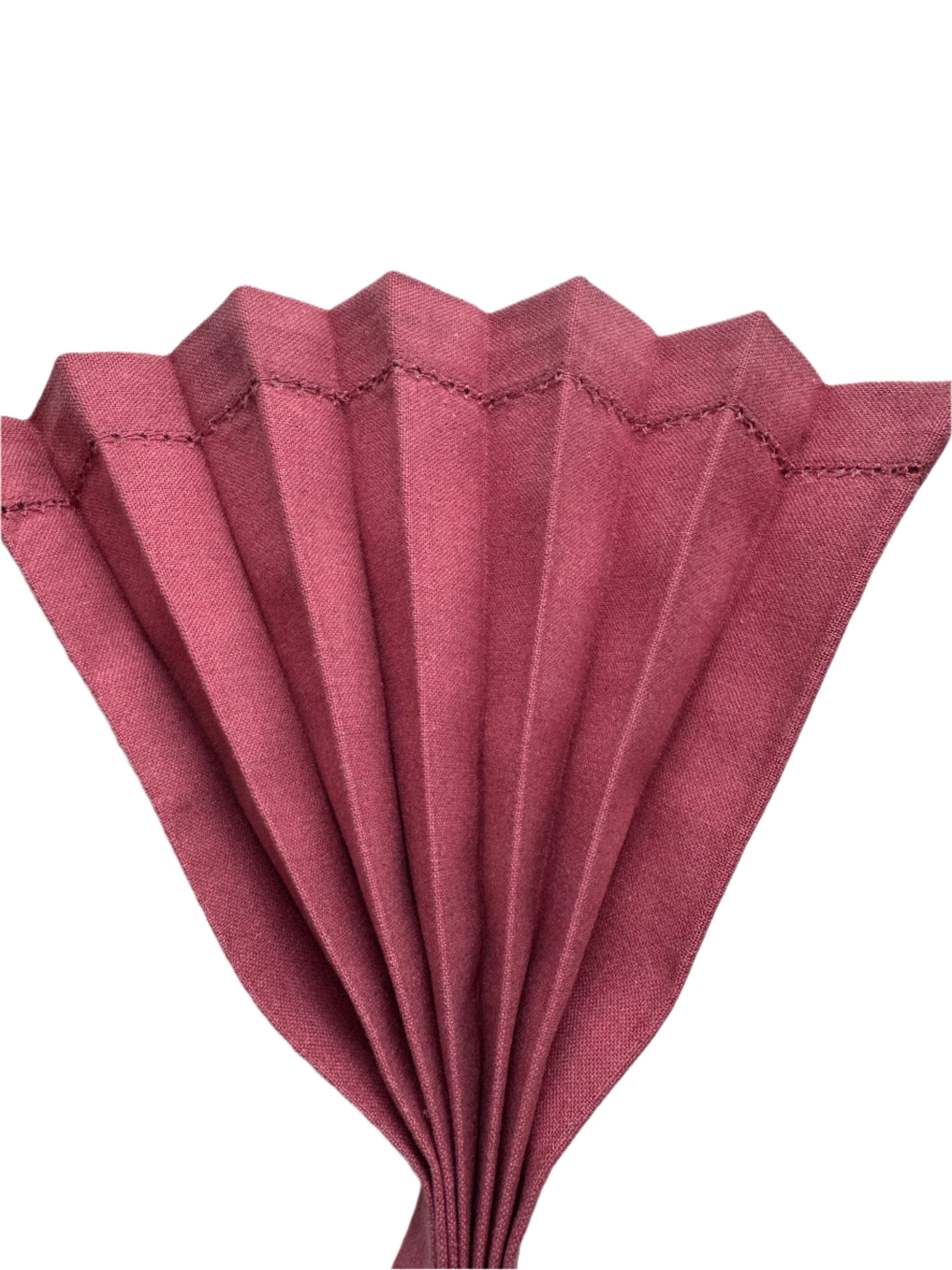 Close-up shot of a fan-folded blush pink cotton napkin, with crisp pleats and neat hemstitch detailing along the edges. The napkin is arranged in an elegant, structured fold, showcasing its fine texture and sophisticated presentation