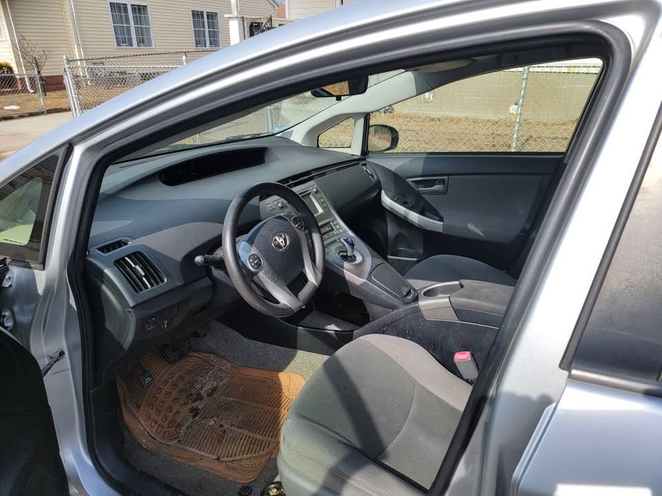 Front interior view of a 2013 Toyota Prius, showing the driver's seat, black and gray cloth upholstery, multifunction steering wheel, center console with gear selector, and dashboard display.