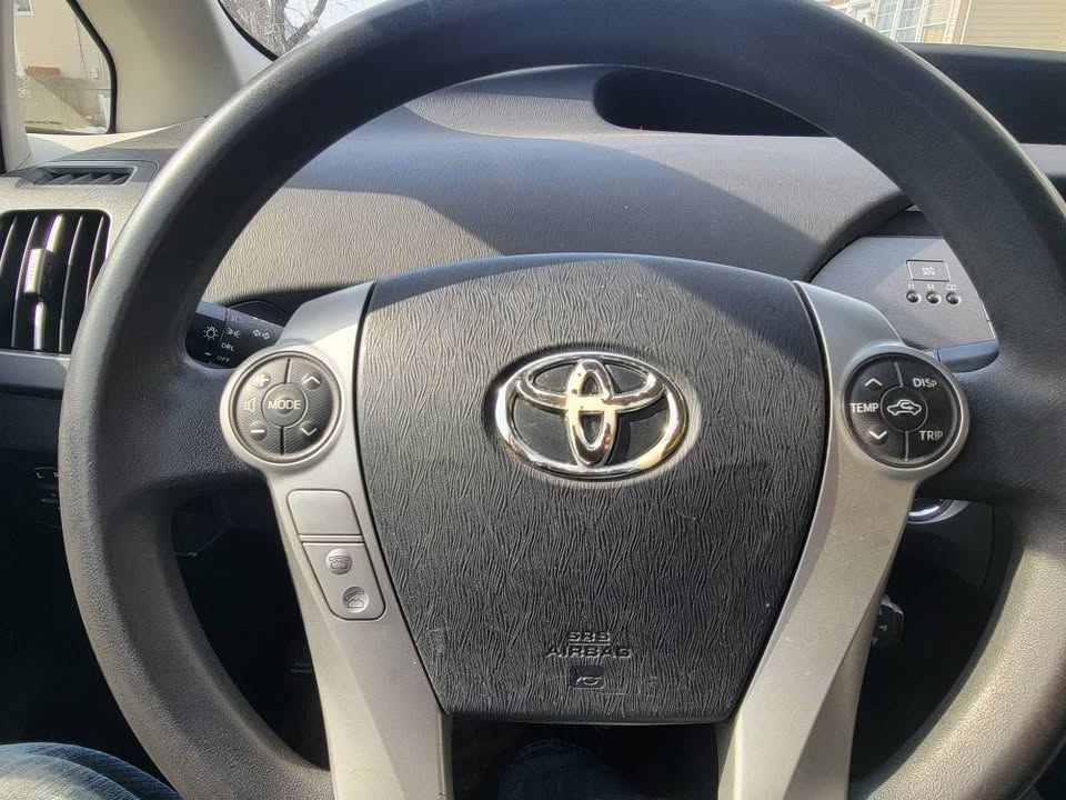 Close-up view of the steering wheel of a 2013 Toyota Prius, featuring the Toyota emblem, textured surface, and multifunction control buttons for audio, temperature, and trip settings.