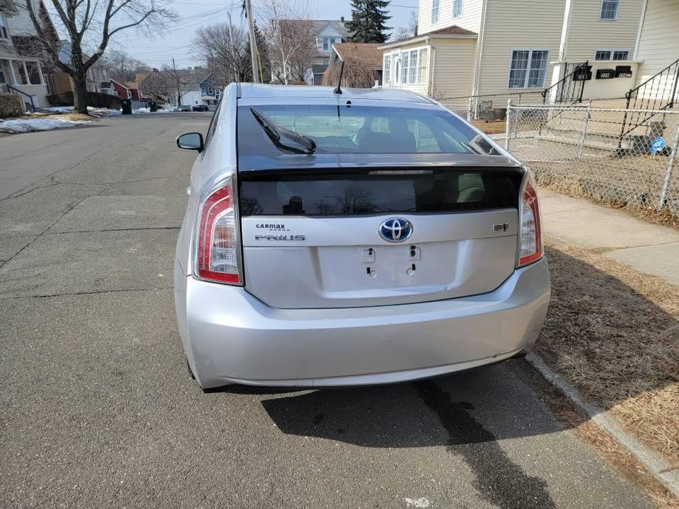Rear view of a silver 2013 Toyota Prius showing the hatchback, tail lights, missing license plate, and CARMAX and Hybrid badges, parked on a residential street.