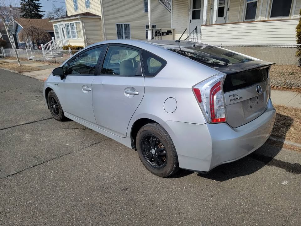 Passenger-side rear angle of a silver 2013 Toyota Prius showing clean body lines, hybrid badge, black wheels, and CARMAX dealership badge, parked in a suburban neighborhood.