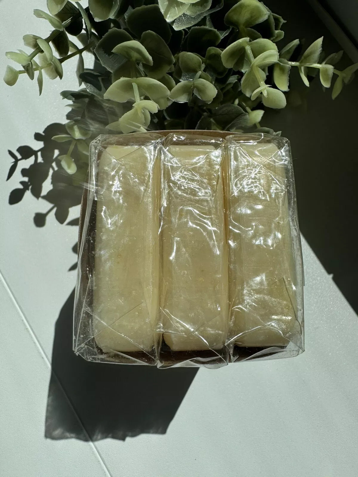 Close-up image of three bars of Ology Oatmeal & Honey soap wrapped in clear plastic, with a soft shadow cast and a leafy plant in the background, set on a white surface