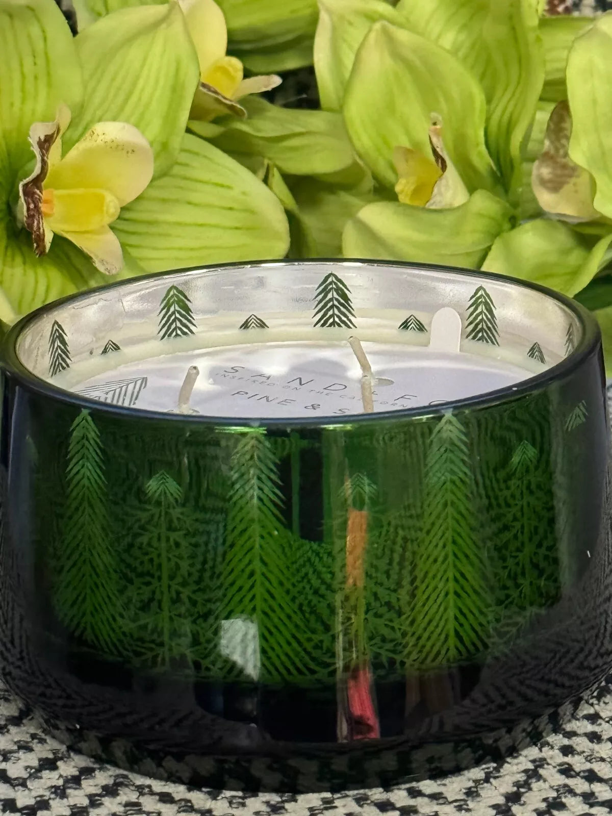 An 18oz Pine and Sage 3-Wick Candle with a Christmas tree motif, hand-poured, displayed in a green glass container surrounded by festive decorations including Christmas trees and wrapped gifts.