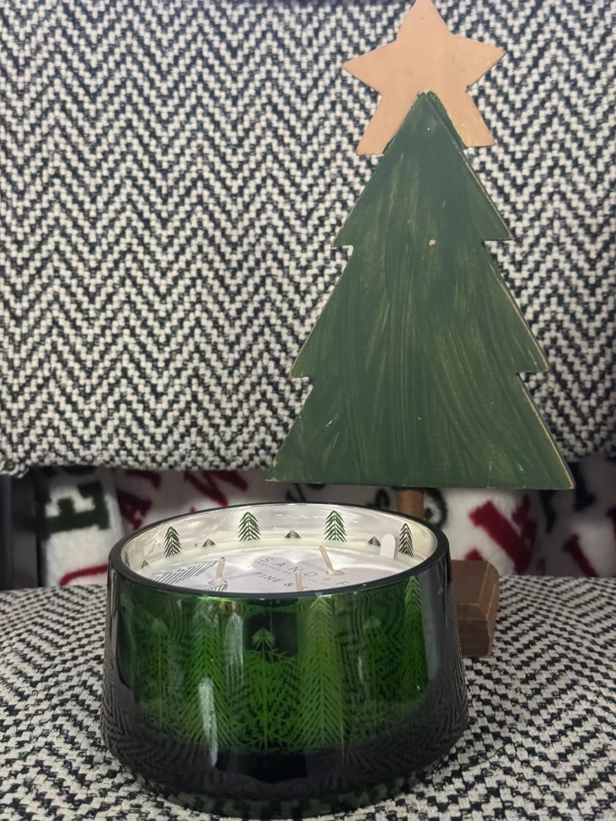 An 18oz Pine and Sage 3-Wick Candle with a Christmas tree motif, hand-poured, displayed in a green glass container surrounded by festive decorations including Christmas trees and wrapped gifts.