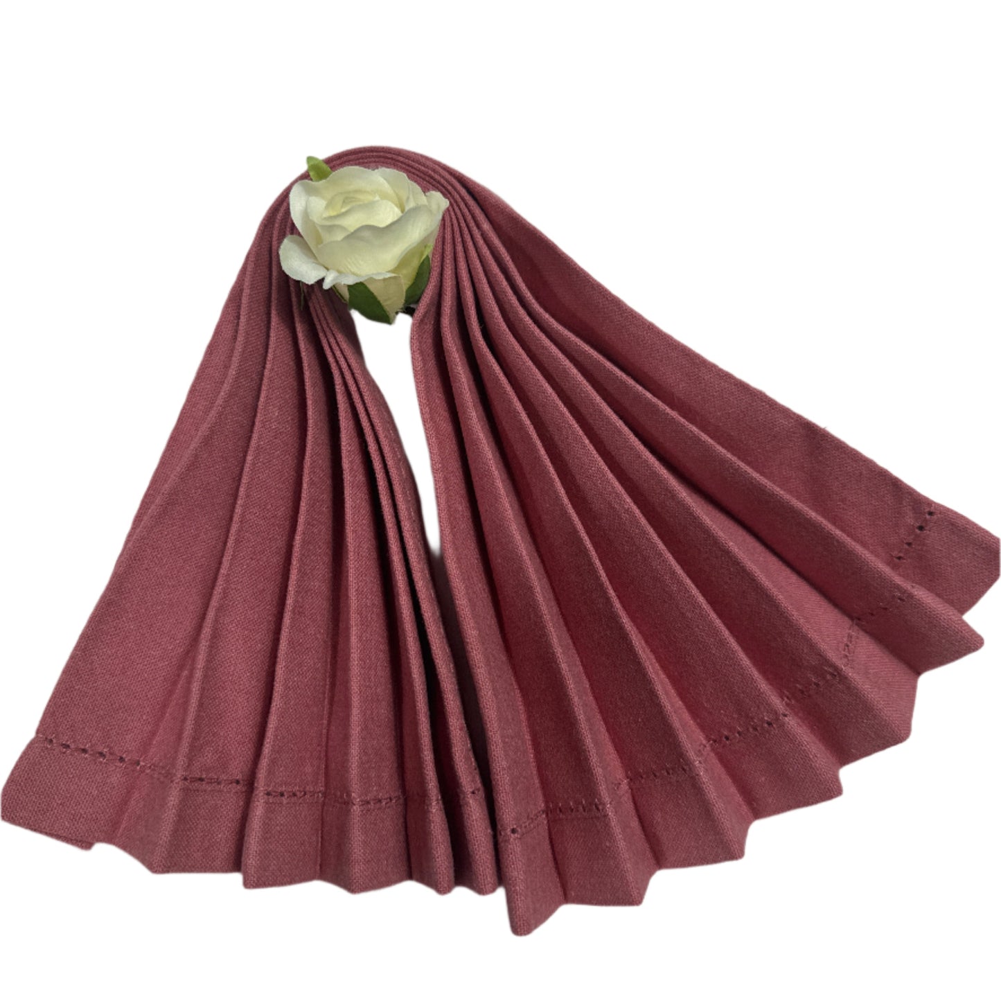 A neatly pleated, rose-colored cotton napkin with delicate hemstitching along the edges, accented by a white artificial rose at the top