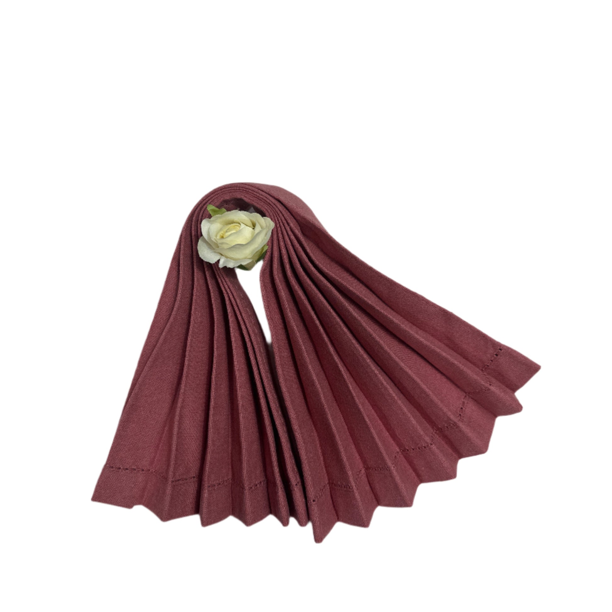 A neatly pleated, rose-colored cotton napkin with delicate hemstitching along the edges, accented by a white artificial rose at the top